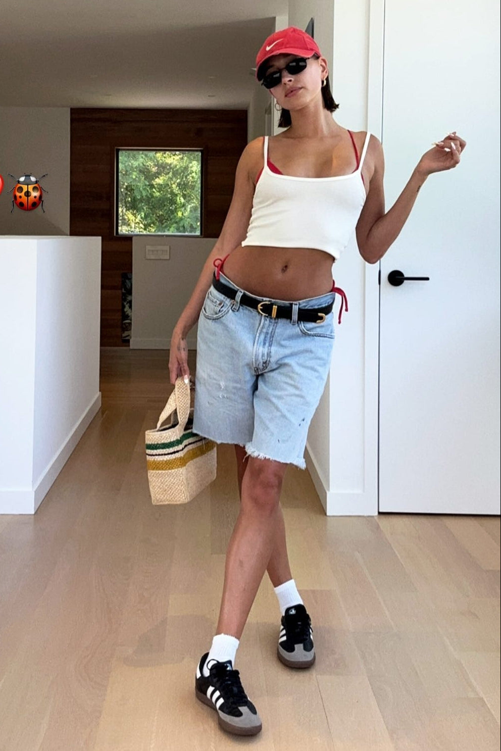 Woman in casual outfit with white tank top, denim shorts, and red cap indoors.