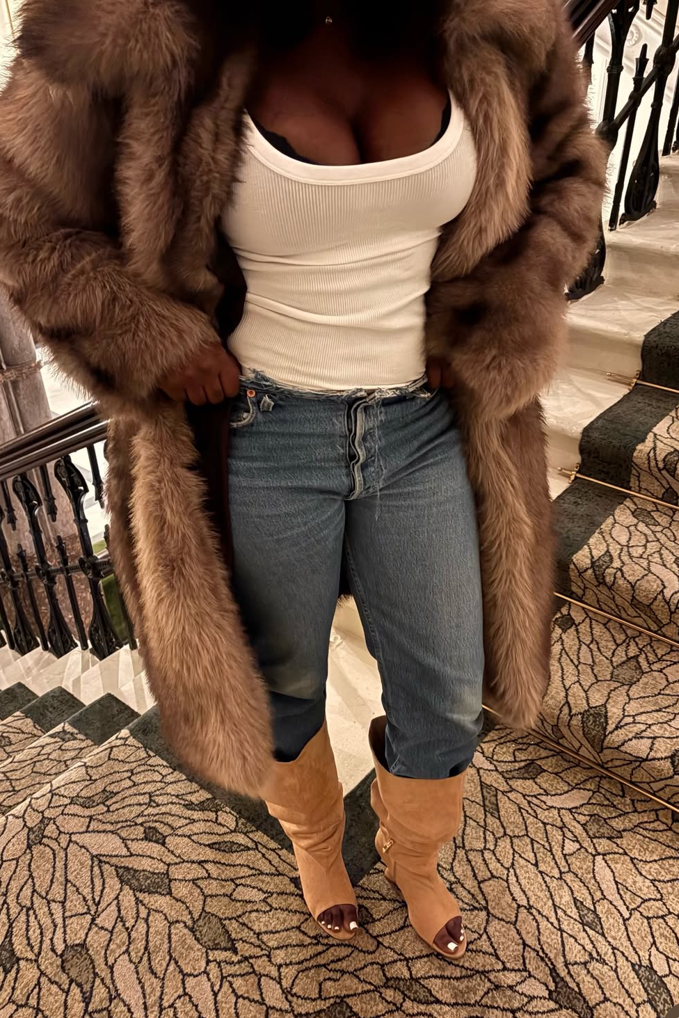 Person wearing a long brown fur coat, white top, blue jeans, and brown boots on a staircase.