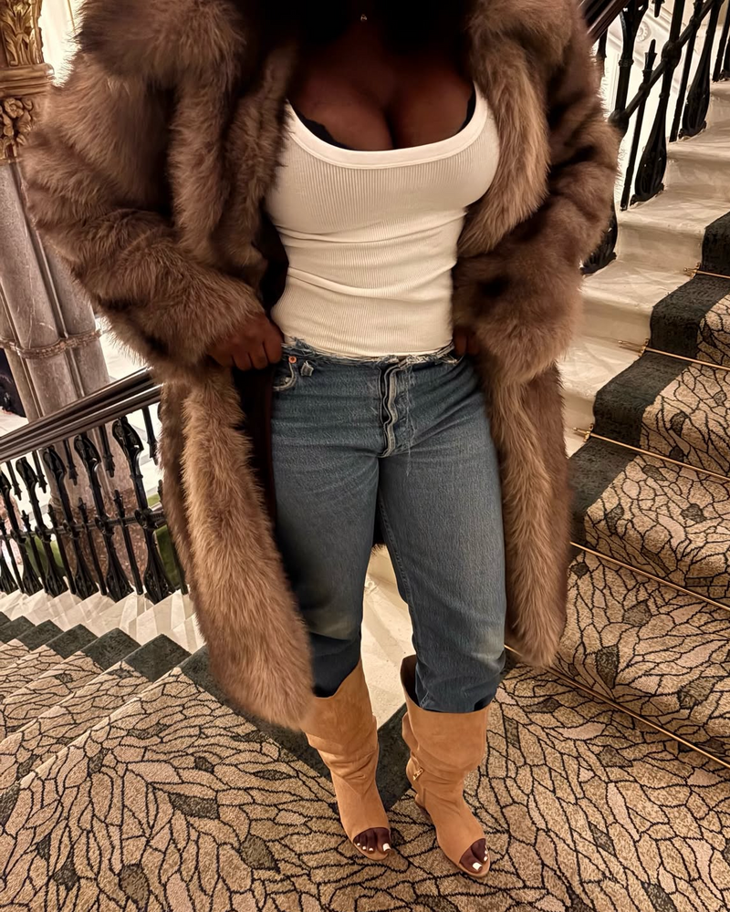 Person wearing a long brown fur coat, white top, blue jeans, and brown boots on a staircase.