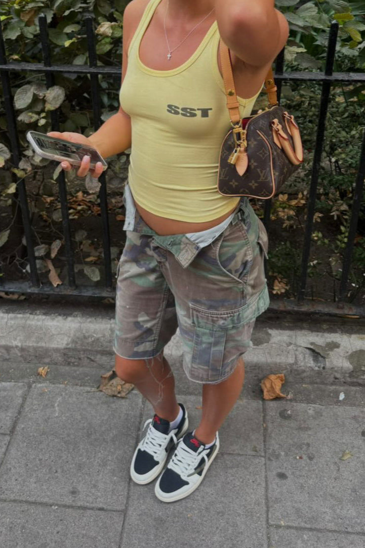 Woman in yellow tank top and camouflage vintage custom shorts standing on a sidewalk with greenery and a brick wall in the background.
