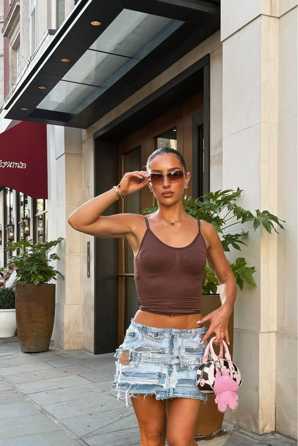 Woman in a brown top and denim shorts standing on a sidewalk with a building in the background.