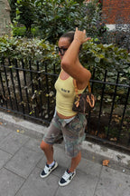 Person wearing a yellow tank top and vintage custom camouflage shorts on a sidewalk with greenery in the background | closet luna